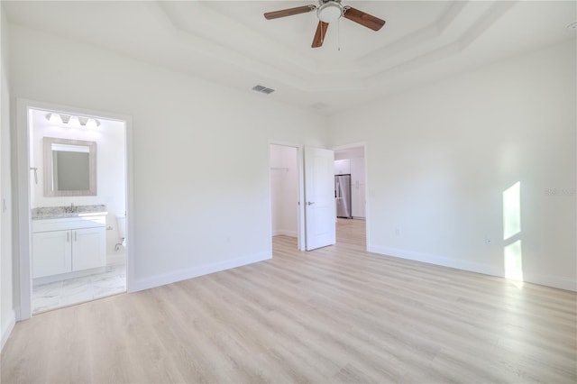 unfurnished bedroom featuring ceiling fan, stainless steel fridge, connected bathroom, a tray ceiling, and light hardwood / wood-style floors