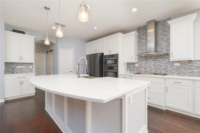 kitchen featuring a barn door, a kitchen island with sink, stainless steel appliances, a sink, and wall chimney exhaust hood