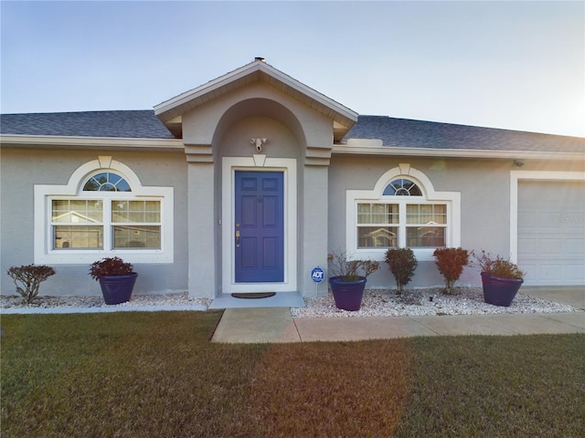 doorway to property featuring a garage, roof with shingles, a lawn, and stucco siding