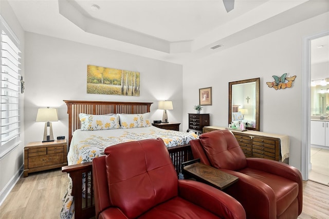 bedroom with a tray ceiling, ensuite bathroom, ceiling fan, and light wood-type flooring