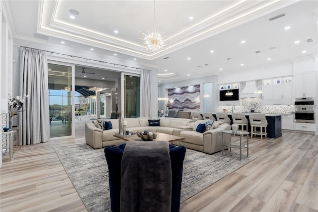 living room with ceiling fan with notable chandelier, ornamental molding, light hardwood / wood-style flooring, and a tray ceiling