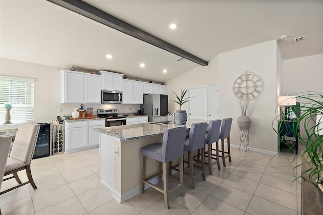 kitchen featuring appliances with stainless steel finishes, lofted ceiling with beams, stone countertops, white cabinetry, and an island with sink