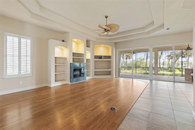 unfurnished living room with built in shelves, ceiling fan, a tile fireplace, plenty of natural light, and a tray ceiling