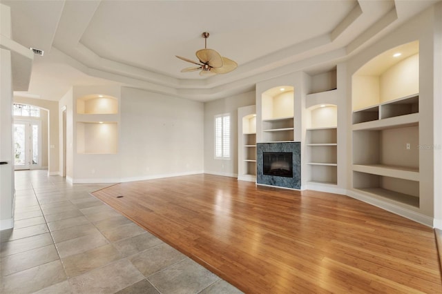 unfurnished living room with a multi sided fireplace, ceiling fan, built in features, light tile patterned floors, and a tray ceiling