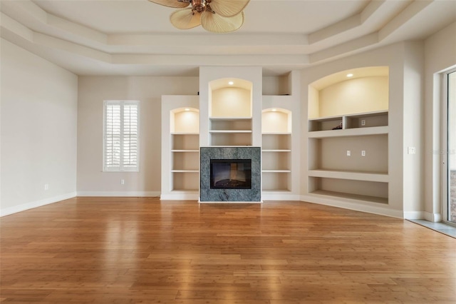 unfurnished living room featuring built in shelves, a raised ceiling, ceiling fan, wood-type flooring, and a fireplace