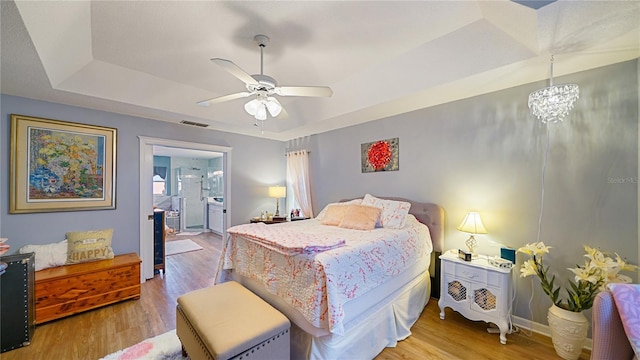 bedroom with ceiling fan with notable chandelier, a tray ceiling, ensuite bath, and light hardwood / wood-style floors