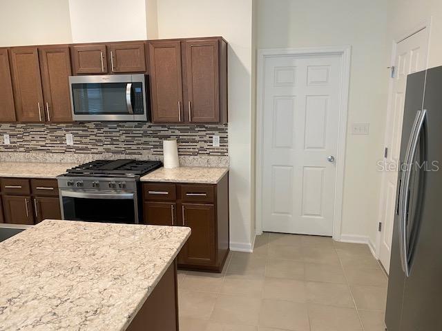 kitchen with stainless steel appliances, light tile patterned floors, light stone counters, decorative backsplash, and dark brown cabinetry