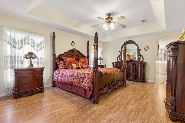 bedroom with ceiling fan, light hardwood / wood-style floors, a raised ceiling, and ensuite bath