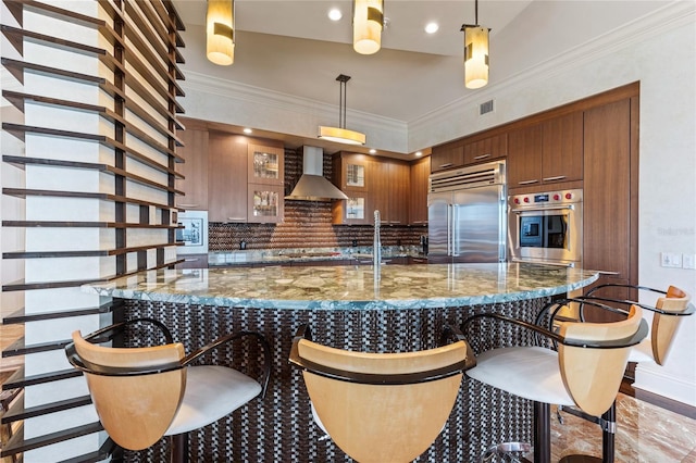 kitchen with a breakfast bar, stone counters, wall chimney range hood, hanging light fixtures, and built in appliances