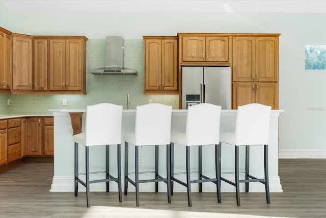 kitchen with dark wood-type flooring, wall chimney range hood, a kitchen breakfast bar, high end refrigerator, and backsplash