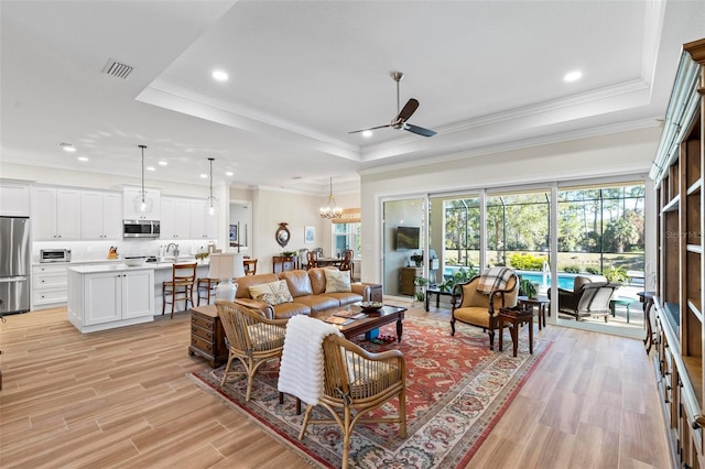 living room with ceiling fan with notable chandelier, ornamental molding, a raised ceiling, and light hardwood / wood-style floors