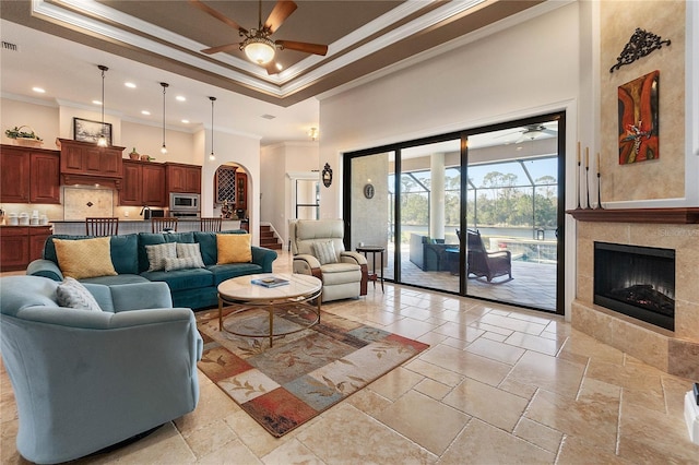 living room with ceiling fan, a raised ceiling, a high ceiling, crown molding, and a tile fireplace