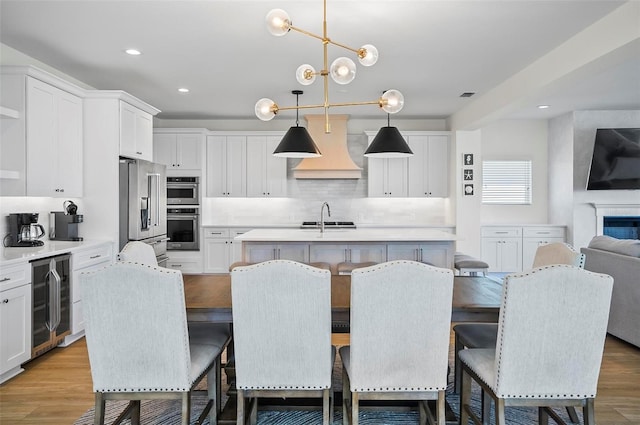 kitchen featuring a breakfast bar, a kitchen island with sink, wine cooler, custom range hood, and decorative light fixtures