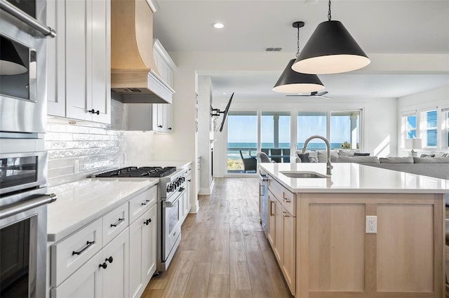 kitchen with pendant lighting, stainless steel stove, premium range hood, white cabinetry, and a center island with sink