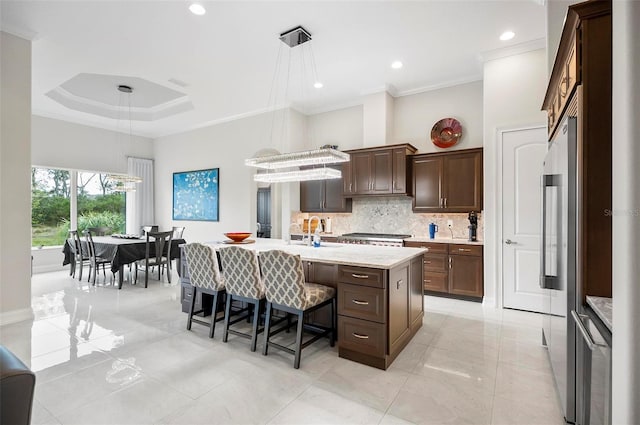 kitchen with hanging light fixtures, ornamental molding, a kitchen island with sink, and a tray ceiling