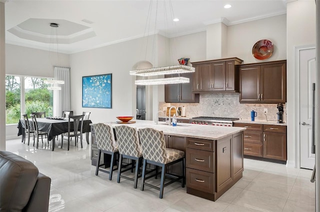 kitchen featuring decorative light fixtures, a kitchen island with sink, a tray ceiling, crown molding, and dark brown cabinets