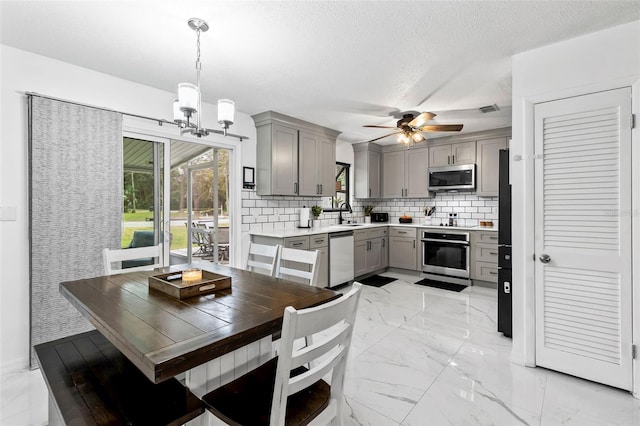 kitchen with pendant lighting, stainless steel appliances, decorative backsplash, gray cabinets, and ceiling fan with notable chandelier