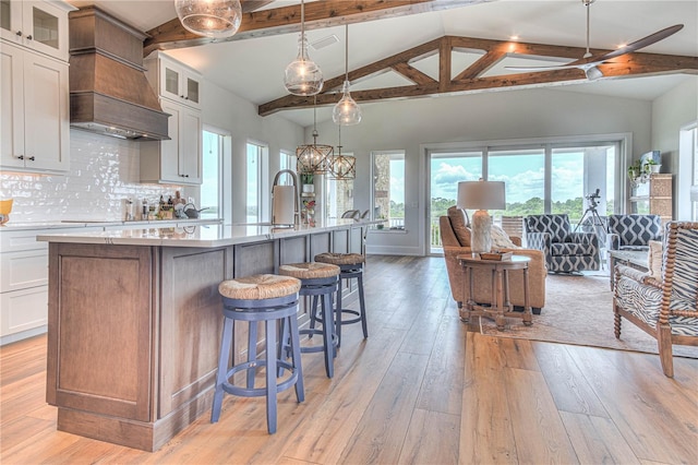 kitchen with decorative backsplash, decorative light fixtures, a kitchen island with sink, vaulted ceiling with beams, and white cabinets