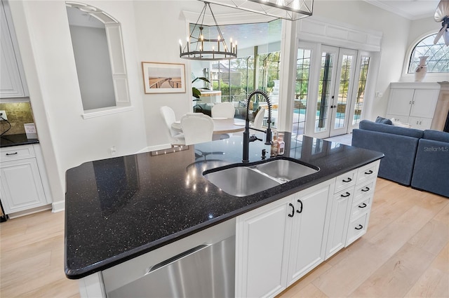 kitchen featuring light hardwood / wood-style floors, stainless steel dishwasher, a center island with sink, an inviting chandelier, and dark stone counters