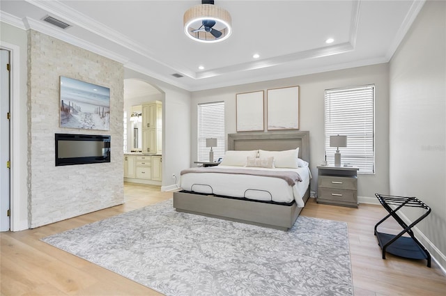 bedroom featuring connected bathroom, a fireplace, a raised ceiling, light hardwood / wood-style flooring, and crown molding