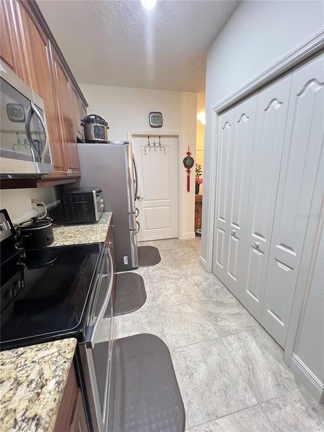 kitchen with a textured ceiling, light stone counters, stainless steel appliances, baseboards, and brown cabinetry