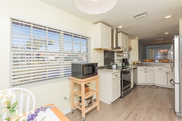 kitchen with appliances with stainless steel finishes, wall chimney range hood, white cabinets, and light wood-type flooring