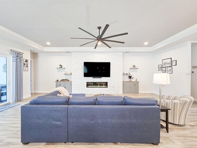 living room with a stone fireplace, crown molding, a textured ceiling, a tray ceiling, and light hardwood / wood-style floors