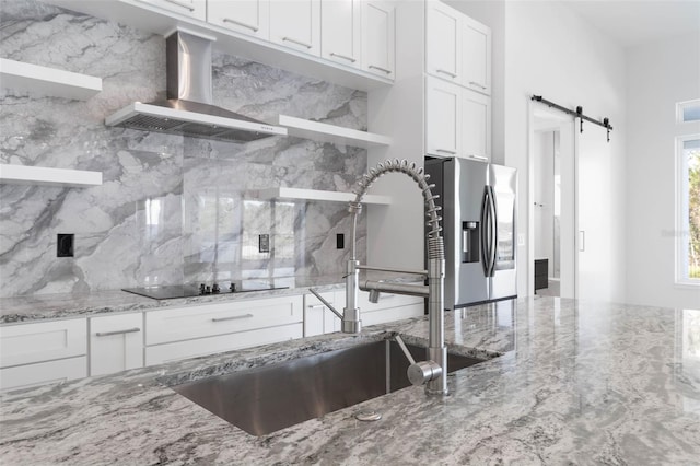 kitchen with white cabinets, stainless steel fridge with ice dispenser, a barn door, black electric cooktop, and wall chimney exhaust hood