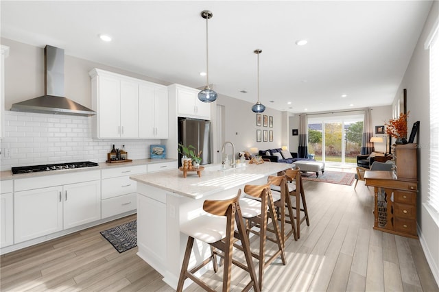 kitchen with pendant lighting, wall chimney range hood, gas cooktop, stainless steel fridge, and an island with sink