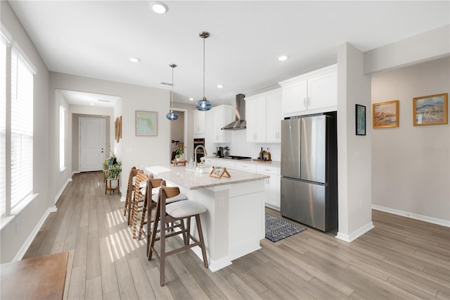 kitchen featuring wall chimney exhaust hood, stainless steel refrigerator, pendant lighting, a kitchen island with sink, and white cabinets