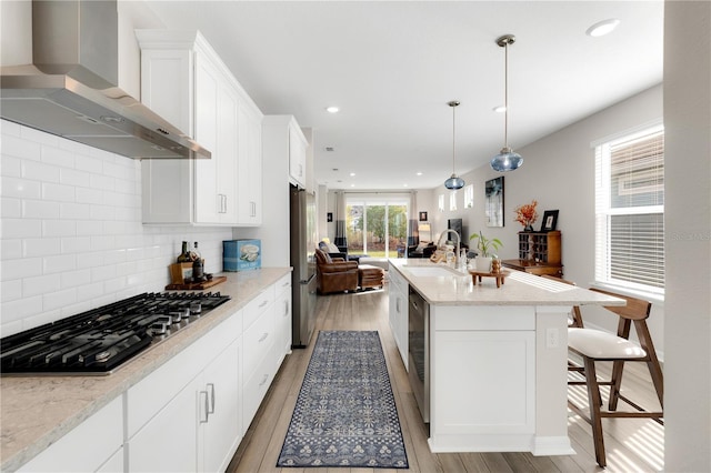 kitchen with sink, a kitchen island with sink, stainless steel appliances, light hardwood / wood-style floors, and wall chimney range hood