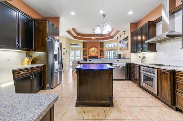 kitchen with appliances with stainless steel finishes, decorative light fixtures, light stone counters, a tray ceiling, and wall chimney exhaust hood