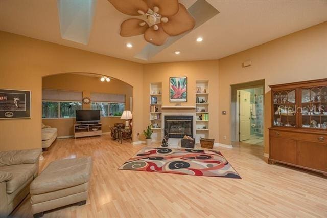 living room featuring built in shelves, ceiling fan, a tray ceiling, and light hardwood / wood-style flooring