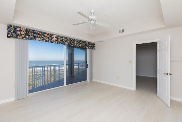 empty room with ceiling fan, a water view, light wood-type flooring, and a tray ceiling