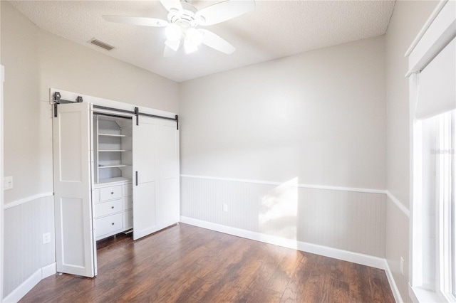 unfurnished bedroom with ceiling fan, a barn door, dark wood-type flooring, and a textured ceiling