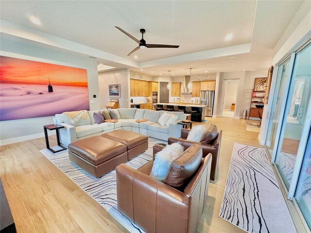 living room with a raised ceiling, ceiling fan, and light wood-type flooring