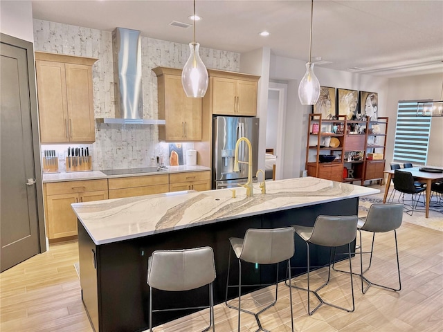 kitchen with light brown cabinetry, an island with sink, black electric stovetop, stainless steel refrigerator with ice dispenser, and wall chimney range hood