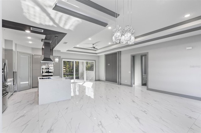 kitchen with a tray ceiling, a kitchen island, pendant lighting, ceiling fan, and wall chimney range hood