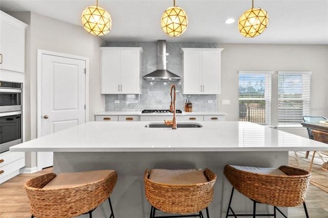 kitchen with stainless steel double oven, light countertops, light wood-style floors, wall chimney exhaust hood, and tasteful backsplash