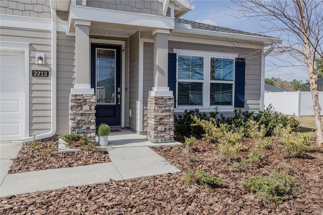 view of exterior entry with stone siding, a shingled roof, an attached garage, and fence