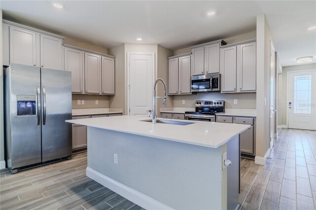 kitchen with wood finish floors, a sink, appliances with stainless steel finishes, gray cabinets, and a center island with sink