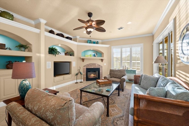 tiled living room with visible vents, a ceiling fan, a glass covered fireplace, crown molding, and built in shelves