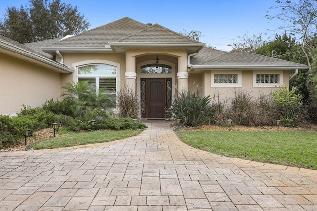 entrance to property with roof with shingles, a yard, and stucco siding