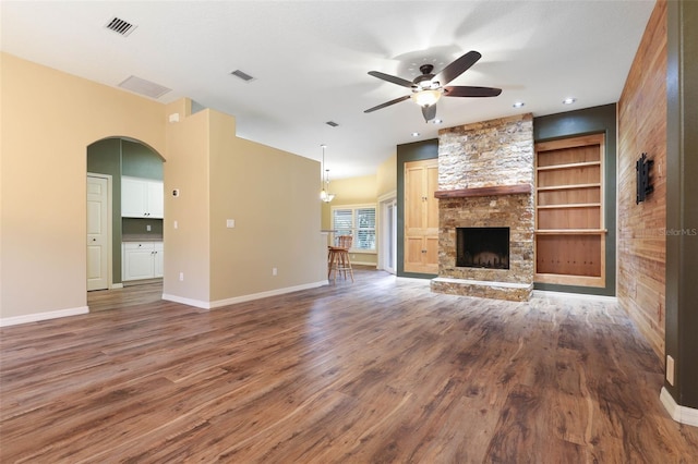 unfurnished living room with a stone fireplace, dark wood-type flooring, built in features, and ceiling fan