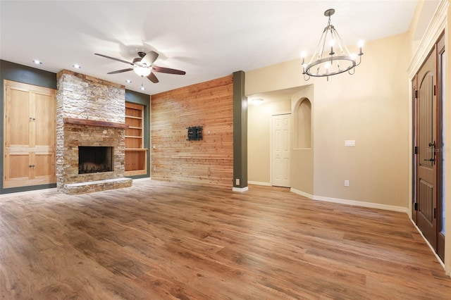 unfurnished living room with a stone fireplace, wooden walls, ceiling fan with notable chandelier, wood finished floors, and built in features