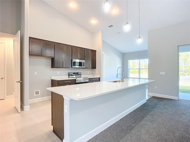 kitchen with sink, a kitchen island with sink, stainless steel appliances, dark brown cabinetry, and decorative light fixtures