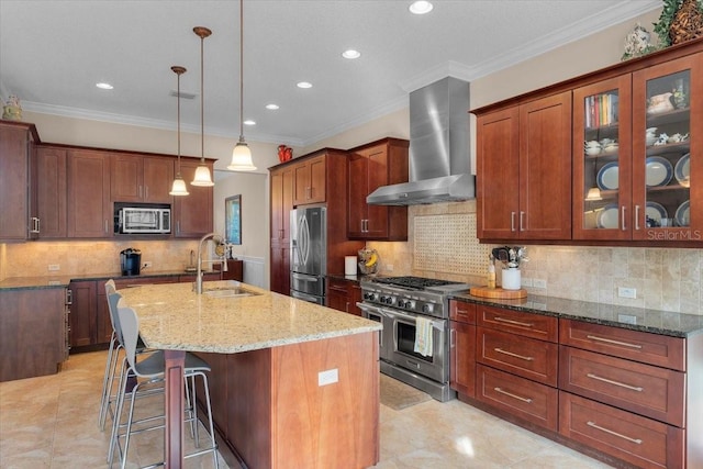 kitchen with appliances with stainless steel finishes, sink, wall chimney range hood, hanging light fixtures, and a center island with sink