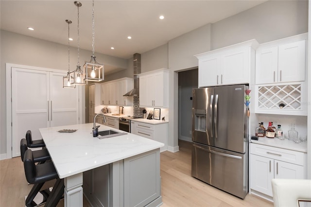 kitchen featuring wall chimney exhaust hood, light wood-style flooring, appliances with stainless steel finishes, a kitchen island with sink, and a sink