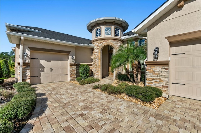 view of exterior entry with a garage, stone siding, decorative driveway, roof with shingles, and stucco siding