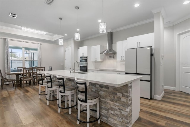 kitchen with pendant lighting, wall chimney range hood, a kitchen island with sink, white refrigerator, and white cabinets